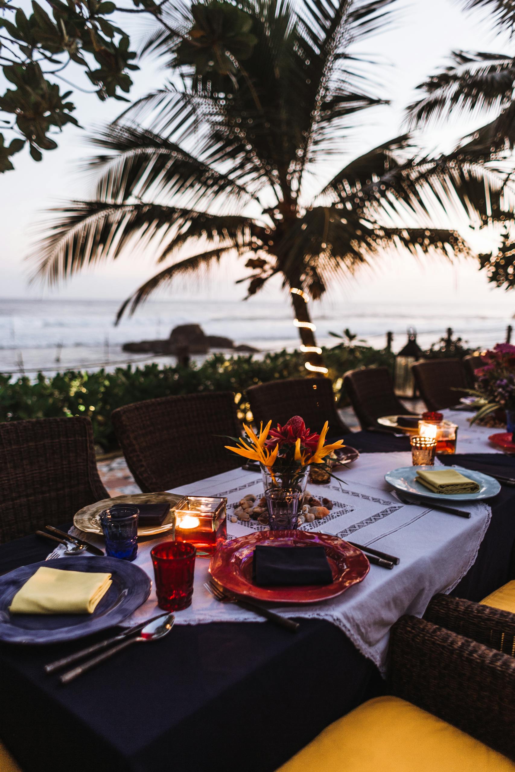 A beautifully set dining table by the seaside with candlelight and vibrant decor at sunset.
