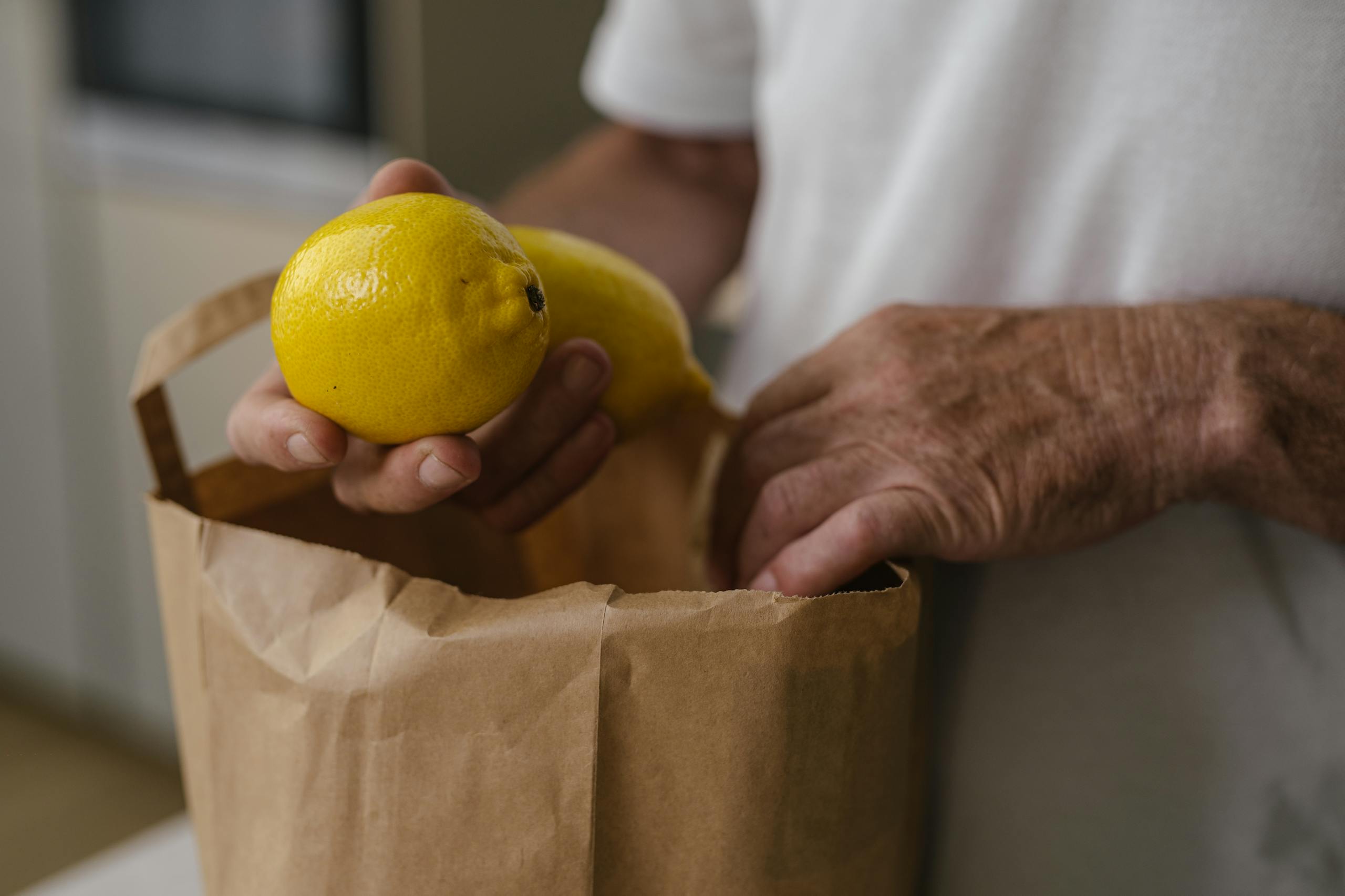 Adult hands placing lemons into a paper bag. Indoor setting with a soft focus background.